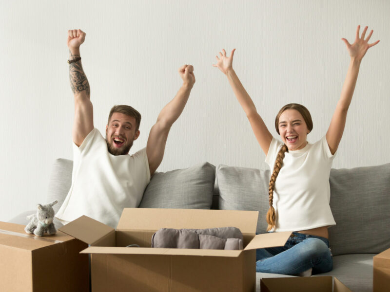 Excited millennial couple moving to new home, happy husband and wife raising hands celebrating apartment purchase, lovers starting living together relocating to first shared flat. Looking at camera